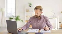 一个留着胡子的中年金发男子在家里的办公室里用笔记本电脑写东西